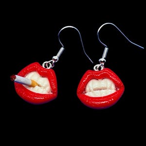 Op de afbeelding: Een paar zilveren oorhangers met rode lippen en witte tanden. Een oorbel heeft een sigaret in de lippen.