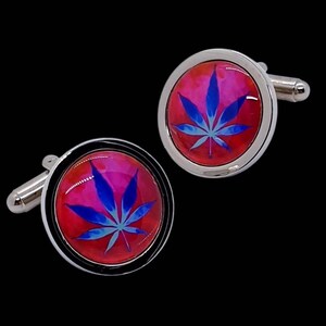 May include: A pair of silver cufflinks with a striking design. Each cufflink showcases a red and pink backdrop with a blue cannabis leaf motif. The cufflinks have a polished, metallic finish and a traditional, circular form.