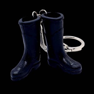 Puede incluir: Un llavero de botas de goma negras en miniatura. El llavero presenta dos pequeñas botas detalladas conectadas por un anillo y una cadena de metal plateado. Las botas tienen un acabado brillante y están sobre un fondo negro.