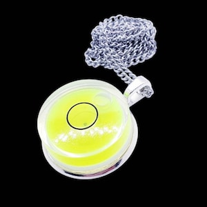 May include: A silver-toned necklace featuring a round pendant with a clear, bubble-like center filled with a bright yellow liquid. A black circle is visible within the liquid, and the pendant is attached to a silver chain. The background is black.