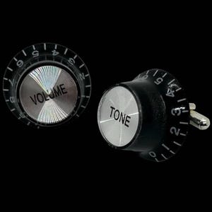 Peut inclure: Deux boutons de manchette, l'un avec une face transparente et le mot "VOLUME" et l'autre avec une face noire et le mot "TONE". Les deux ont un centre argenté et des chiffres noirs sur le bord.