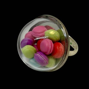 Könnte beinhalten: Eine transparente Teetasse aus Glas, gefüllt mit bunten Makronen. Die Makronen sind in den Farbtönen Rosa, Rot, Lila und Grün gehalten. Die Tasse hat einen Henkel und steht vor einem schwarzen Hintergrund.