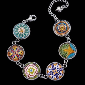 May include: A silver bracelet with seven circular charms, each displaying a vibrant mosaic pattern. The charms are linked by small silver chains and a heart-shaped charm. The mosaic designs feature blue, green, yellow, red, and orange hues, creating a colourful accessory.