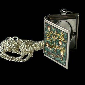 May include: A silver-toned locket with a chain, featuring a hinged design. The locket's cover displays the title "Little Women" in gold script, surrounded by illustrations of women and floral elements. The chain has a detailed, textured design.