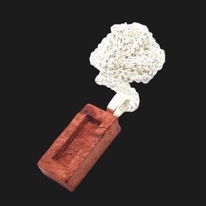 Puede incluir: Un colgante con un pequeño dije de ladrillo rectangular en tonos marrón rojizo, con un detalle rectangular empotrado. El ladrillo está conectado a un diseño plateado en forma de nube. El colgante cuelga sobre un fondo negro.