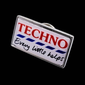 Op de afbeelding: Een rechthoekige speld met een zilveren rand. De speld toont het woord "TECHNO" in rode letters boven blauwe strepen, met de zin "Every little helps" in zwarte letters eronder.