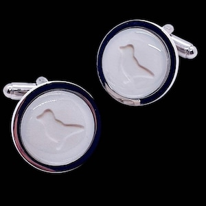 Puede incluir: Un par de gemelos plateados con un fondo blanco y una silueta de pájaro. Los gemelos tienen un borde azul oscuro y un acabado plateado brillante. El diseño del pájaro está en el centro de cada gemelo.