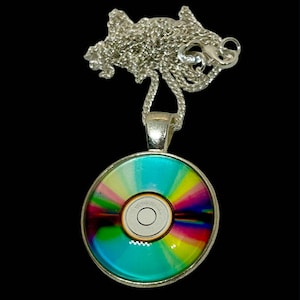 Może przedstawiać: Srebrny naszyjnik z okrągłą zawieszką z kolorowym, tęczowym wzorem. Zawieszka przypomina płytę CD, z centralnym białym okręgiem i żywym spektrum kolorów promieniujących na zewnątrz. Łańcuszek również jest srebrny.