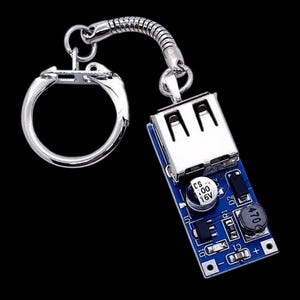 Może przedstawiać: Srebrny brelok do kluczy z portem USB i niebieską płytką drukowaną. Brelok ma srebrny pierścień i elastyczny, spiralny łącznik. Płytka drukowana ma kilka małych elementów, w tym kondensator.
