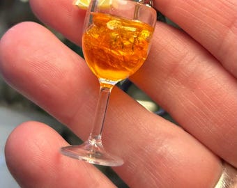 May include: Miniature cocktail charm in a clear glass with orange liquid and ice cubes, topped with a lemon slice. The charm is held between fingers, showcasing the delicate details of the drink.