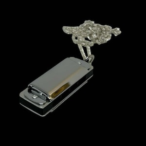 May include: A silver-colored harmonica pendant with a decorative top. The harmonica has a shiny, metallic finish and black accents. The pendant is suspended from a silver chain, and the decorative top has a detailed, textured design. The background is black.