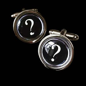 Puede incluir: Un par de gemelos plateados con fondo negro. Cada gemelo presenta un diseño circular con un signo de interrogación blanco en el centro. Los gemelos tienen un acabado metálico brillante.