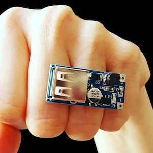 Pode incluir: Uma pequena placa de circuito azul retangular com uma porta USB prateada e vários componentes eletrônicos. A placa é segurada em uma mão, mostrando seu tamanho compacto e design. Os componentes incluem um capacitor preto e outras peças pequenas.
