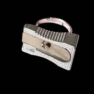 Puede incluir: Un anillo plateado con un diseño de sacapuntas en miniatura. El anillo tiene una banda ajustable y una réplica detallada de un sacapuntas, con una cuchilla y detalles de tornillos. El fondo es negro liso.