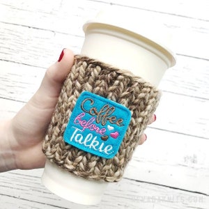 May include: A knitted coffee cup sleeve in shades of brown and tan, with a turquoise square patch. The patch features the embroidered phrase "Coffee before Talkie" in brown and pink, with heart accents. The sleeve is designed to fit a standard coffee cup.