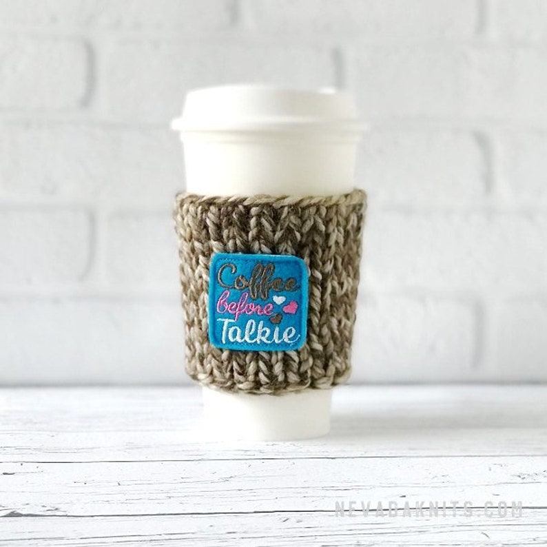 May include: A white disposable coffee cup with a knitted brown and beige coffee sleeve. The sleeve has a blue square patch with the text "Coffee Before Talkie" in pink and white. The cup is on a white wooden surface.