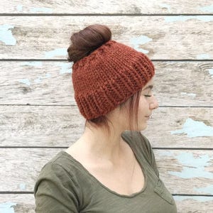 May include: A woman with a messy bun wears a rust-colored knitted beanie with a fold-over brim. The beanie is made of a soft, chunky yarn. The woman is standing in front of a weathered wooden wall.
