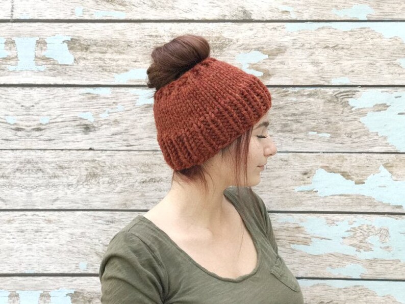 May include: A woman with a messy bun wears a rust-colored knitted beanie with a fold-over brim. The beanie is made of a soft, chunky yarn. The woman is standing in front of a weathered wooden wall.