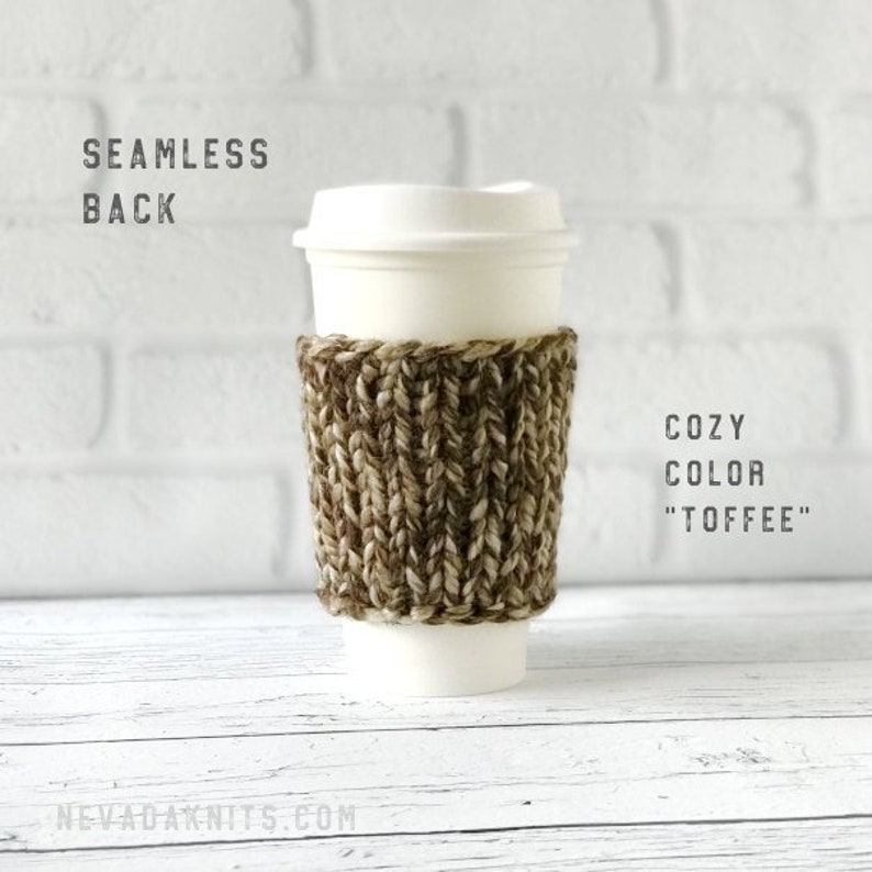 May include: A white paper coffee cup with a brown and tan knitted cozy. The cozy is wrapped around the cup and has a textured appearance. The text "COZY COLOR 'TOFFEE'" is visible. The background is a white brick wall and a white wooden surface.