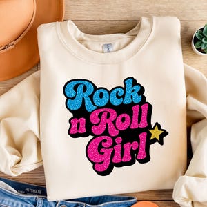 May include: Cream-colored sweatshirt with the text "Rock n Roll Girl" in blue and pink glitter lettering. A yellow glitter star is next to the word "Girl". The sweatshirt is on a wooden surface with a brown hat and jeans in the background.