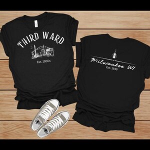 Könnte beinhalten: Zwei schwarze T-Shirts mit weißen Grafiken und ein Paar graue Turnschuhe. Ein Shirt trägt die Aufschrift "THIRD WARD" mit einer Illustration und "Est. 1850s". Das andere trägt die Aufschrift "Milwaukee WI" mit einer Flaschengrafik und "Est. 1846".