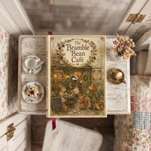 May include: A hardcover book titled The Bramble Bean Café with an illustration of anthropomorphic animals at a cafe. Beside the book are two small plates, one with a tiny cup and spoon, the other with a miniature muffin, and a small candle holder.