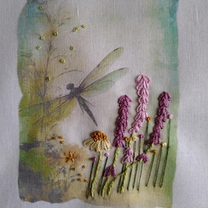 May include: Embroidered artwork depicting a dragonfly and wildflowers. The design includes a dragonfly with delicate wings, lavender-coloured flowers, and yellow-centred daisies. The background has soft green and blue hues, creating a serene, nature-inspired scene.