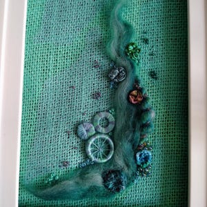 May include: A framed textile art piece featuring a textured mint green hessian background adorned with a flowing wave of teal wool roving, embroidered circular elements, small dark beads, and various buttons in shades of blue and green.