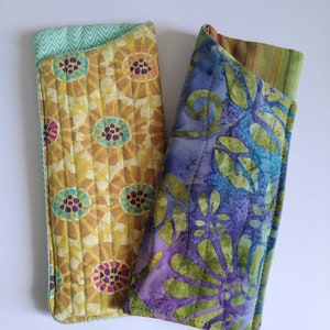 Puede incluir: Dos estuches para gafas de tela, uno con un patrón floral amarillo y verde y el otro con un patrón floral azul y verde.