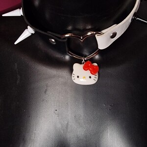 Può includere: Un collare in pelle nero e bianco con punte argentate e un ciondolo a forma di cuore con un personaggio di Hello Kitty con un fiocco rosso.