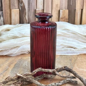 Könnte beinhalten: Eine tief burgunderrote Glasvase mit gerippter Textur. Die Vase steht auf einer Holzoberfläche mit einem Stück Treibholz davor.