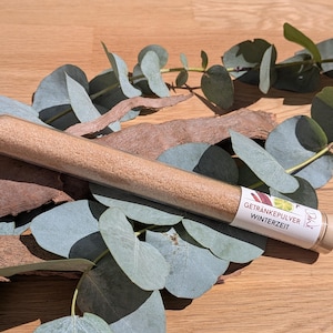 Op de afbeelding: Een heldere glazen buis gevuld met lichtbruin poeder, met het label "Getränkepulver Winterzeit". De buis rust op een bed van eucalyptusbladeren en stukken hout, tegen een houten oppervlak. Het label heeft een kleurrijk ontwerp.