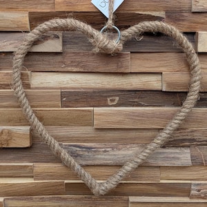 May include: A heart-shaped decoration crafted from thick, natural rope. The rope is knotted at the top and held by a metal ring. The backdrop is a wooden wall with various shades of brown.