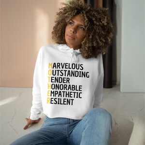 May include: White cropped hoodie with a black and gold graphic that reads "MOTHER" with the letters spelled out as an acronym for "Marvelous, Outstanding, Tender, Honorable, Empathetic, Resilient".