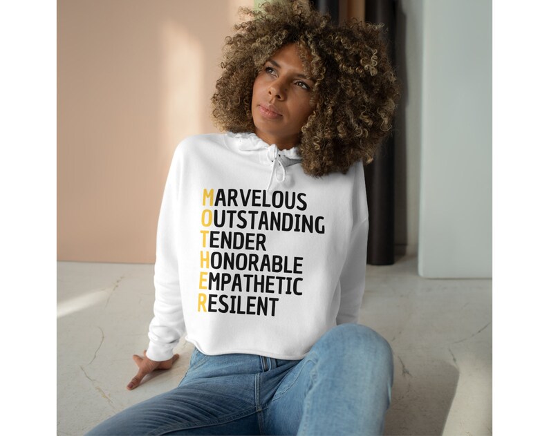 May include: White cropped hoodie with a black and gold graphic that reads "MOTHER" with the letters spelled out as an acronym for "Marvelous, Outstanding, Tender, Honorable, Empathetic, Resilient".