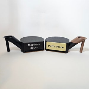 May include: Two black and gray ashtrays with angled stems and rounded platforms. One has the text "Martha's House" and the other has "Puff's Place" on a gold-colored label. The ashtrays are on a white surface.