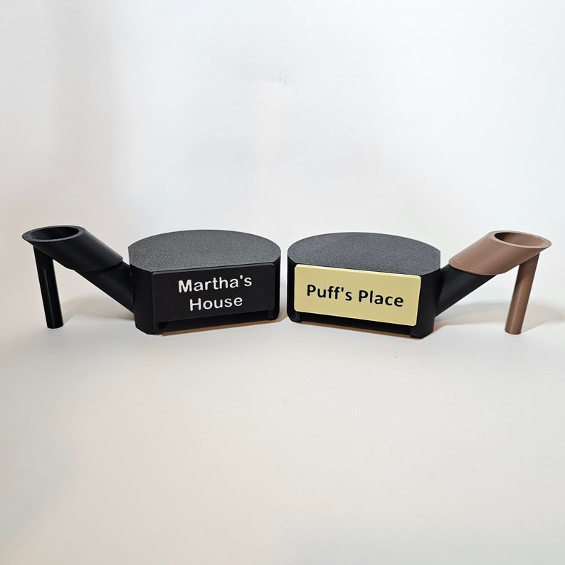 May include: Two black and gray ashtrays with angled stems and rounded platforms. One has the text "Martha's House" and the other has "Puff's Place" on a gold-colored label. The ashtrays are on a white surface.