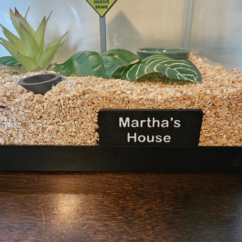 May include: A terrarium with a sign that reads "Martha's House". The terrarium contains wood shavings, artificial plants, and a small ceramic bowl. A sign warns of a "vicious snake".