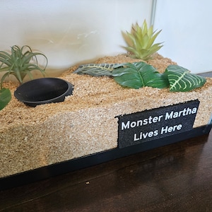 May include: A small terrarium with a black plastic bowl, wood shavings, and artificial plants. A black sign reads "Monster Martha Lives Here".