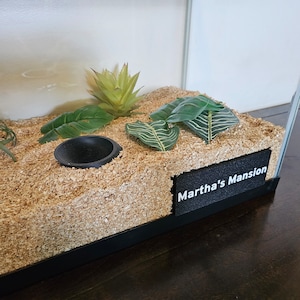 Puede incluir: Un pequeño terrario con un cuenco de plástico negro, virutas de madera y tres plantas verdes. El terrario tiene una etiqueta negra en la parte delantera que dice "Martha's Mansion".