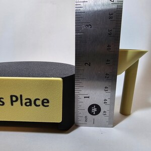 May include: A black and yellow sign with the text "s Place" is next to a black object. A yellow table leg is next to the black object. A ruler shows the table leg is approximately 1.5 inches tall.