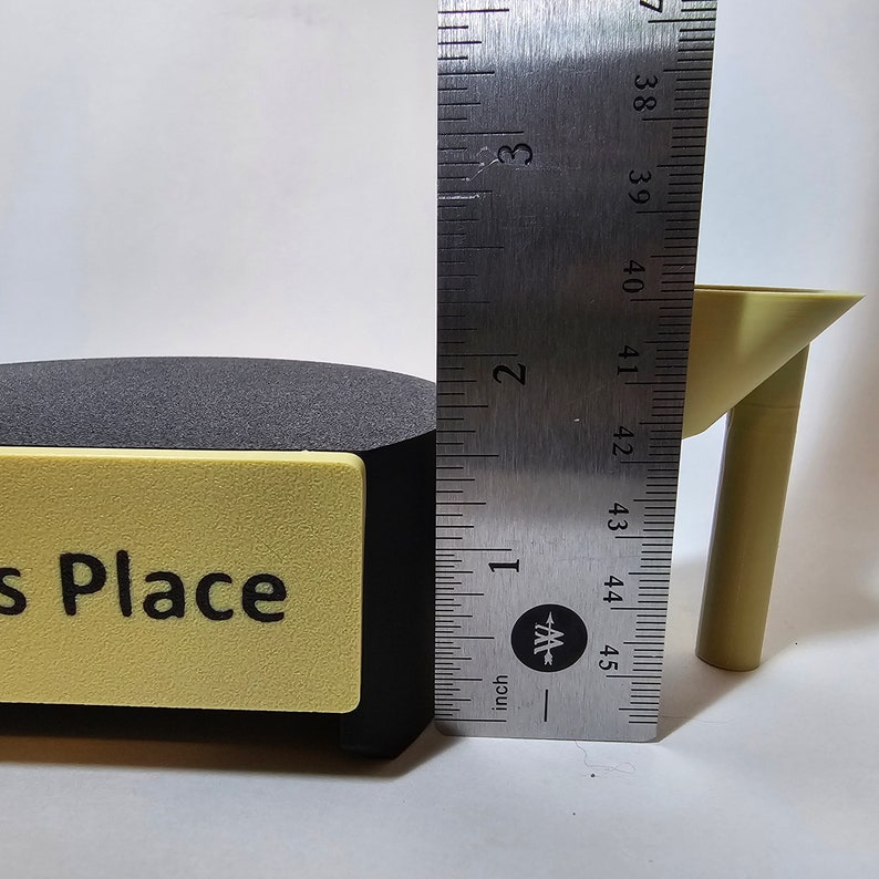 May include: A black and yellow sign with the text "s Place" is next to a black object. A yellow table leg is next to the black object. A ruler shows the table leg is approximately 1.5 inches tall.