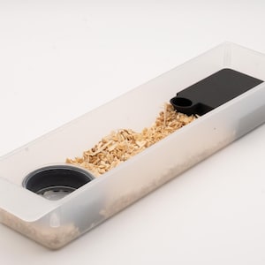 May include: A rectangular, translucent plastic tray filled with light-coloured wood shavings. The tray contains a circular black bowl insert on one end and a flat, black rectangular component with a small circular opening on the other end.