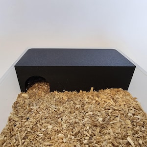 Puede incluir: Escondite de plástico negro para animales pequeños, como hámsters o jerbos. El escondite tiene una sola abertura y se coloca sobre una cama de virutas de madera.
