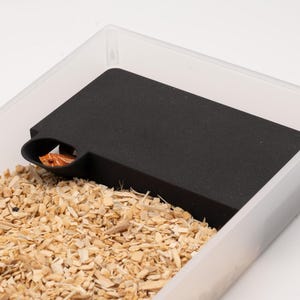 May include: A clear plastic rectangular enclosure containing a black rectangular hide box with a circular opening. Inside the hide box, a small orange and white patterned snake is visible. The enclosure is filled with light brown wood shavings.