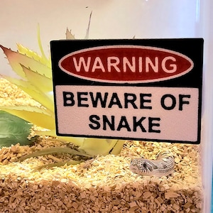 May include: A warning sign that reads "WARNING BEWARE OF SNAKE" in a black frame with a red oval that says "WARNING" inside. A white snake is visible in the bottom right corner of the image.