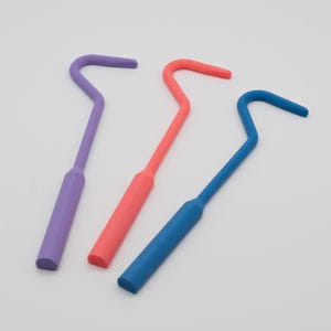 May include: Three tick removal tools in purple, coral, and blue. Each tool has a handle and a curved hook at the end, designed for safely removing ticks. The tools are made of a solid, smooth material.