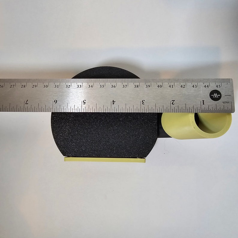 May include: A black, circular object with a yellow rectangular base and a yellow cylindrical tube attached. A metal ruler is placed across the object, measuring approximately 5 inches in width.