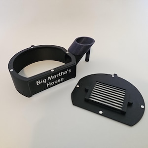 Könnte beinhalten: Ein schwarzes 3D-gedrucktes Objekt mit dem Text "Big Martha's House". Das Objekt hat eine gebogene Basis, einen zylindrischen Halter und ein separates, geschlitztes Teil. Weiße Punkte sind auf dem Objekt sichtbar.