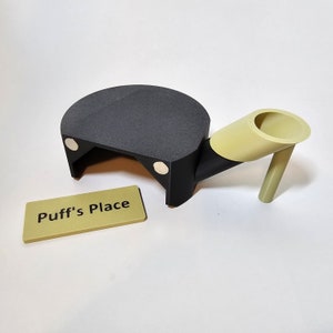May include: A black and tan object with a semi-circular top and a cylindrical extension. The object has a small rectangular sign that reads "Puff's Place". The object is likely a stand or holder.